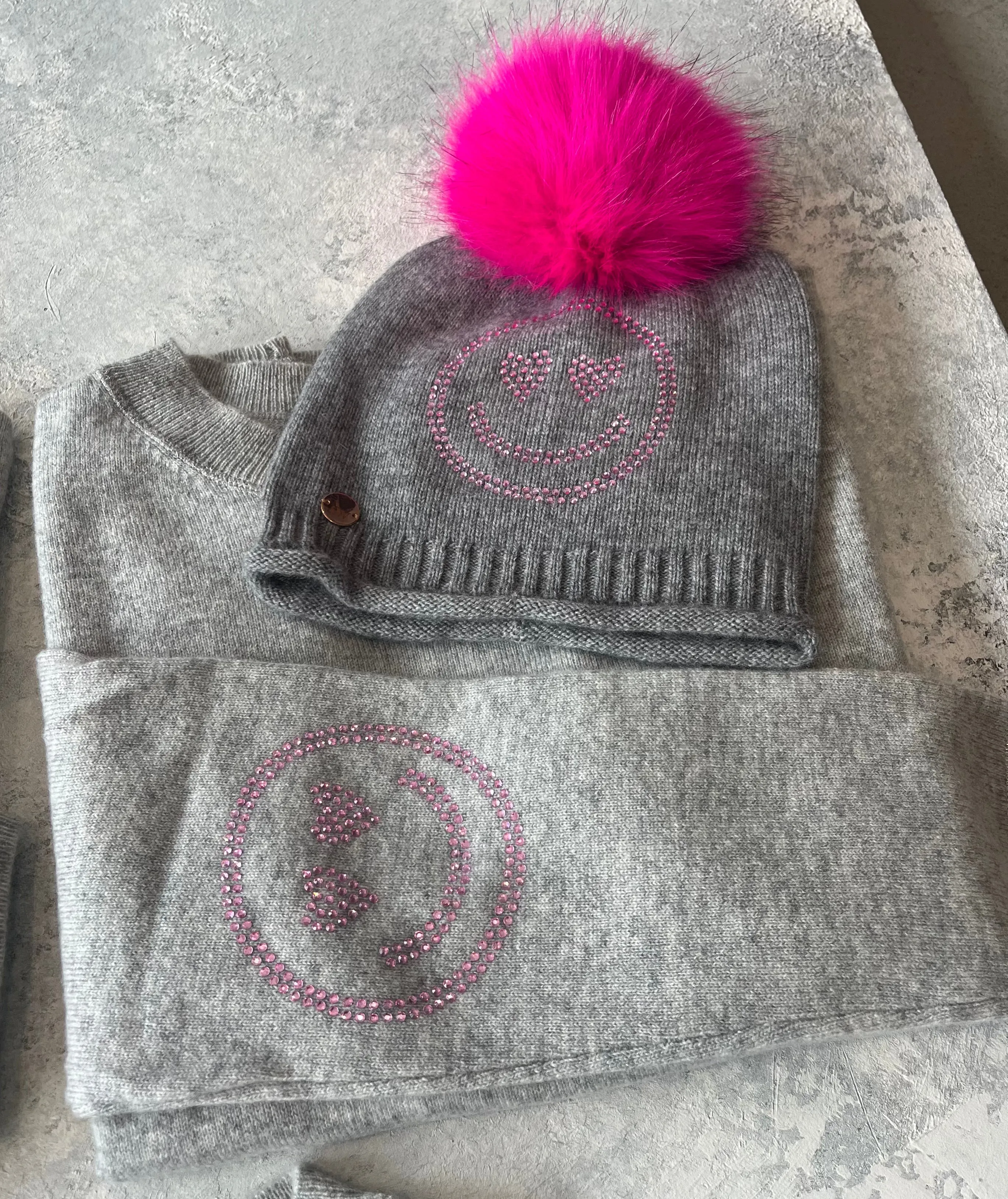 Grey cashmere plain knit beanie with a pink smiley embellishment & faux fur pom